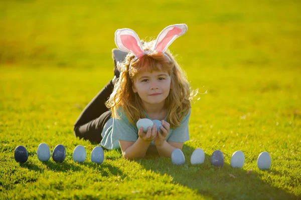 孩子们猎取复活节彩蛋。孩子们正在收集复活节彩蛋，躺在草地上。复活节快乐贺卡。带着复活节彩蛋和野兔耳朵的小男孩. — 图库照片