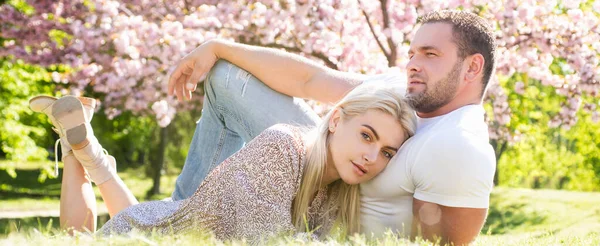 Spring couple in love, banner. Couple relaxing on grass in blossom park. Valentines day concept. Spring couple. Outdoor portrait of young lovers couple near sakura. — Stock Photo, Image