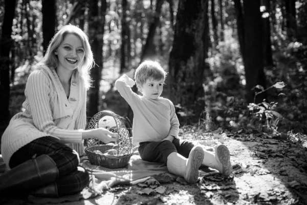 Young mother with sun sit on the picnic blanket at sunny autumn forest and having amazing time. Busket for picnic with food stuff. Smiling happy mum and baby boy at autumn fall picnic. — Fotografia de Stock