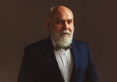 Gülümseyen ve kendinden emin, kel, kıdemli bir iş adamının portresi, takım elbise ve kravat takıyor, gri bir duvarın önünde..
