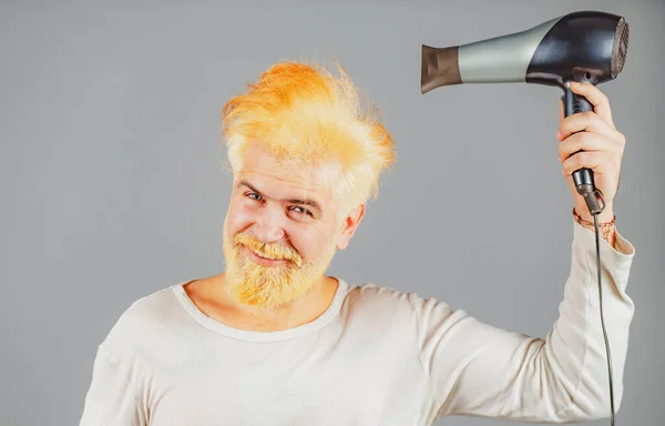 Красивый рыжий мужчина с длинными волосами сушит волосы феном. Блондин с феном. — стоковое фото