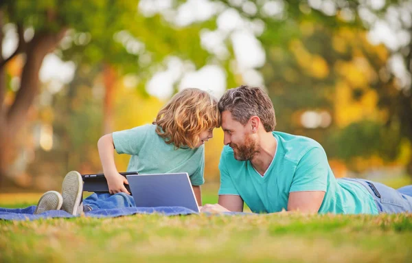 Padre feliz con el uso de su hijo portátil. Papá y el niño de la escuela mirando la pantalla y la tableta, viendo video lección, sentado en la hierba. Fin de semana familiar. Videollamada de un nieto a los abuelos. — Foto de Stock