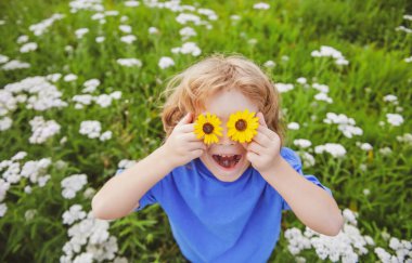 Komik Çocuk ve Papatyalar. Çiçekli, mutlu, sarı saçlı çocuk. Gözleri papatya çiçeğiyle çimlerin üzerinde uzanıyor. Papatya gözlü çocuk, yeşil çimlerin üzerinde yaz parkında.