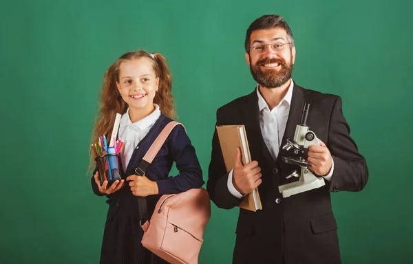 Шкільна дівчина зі смішним здивованим вчителем у школі. Студійний портрет репетитора та молодої школярки з рюкзаком та підручником на зеленому фоні дошки . — стокове фото