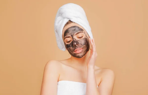 Mujer con máscara de carbón tocándose la nariz. Máscara facial de barro, máscara de barro facial spa. Hermosa mujer con procedimiento facial de barro cosmético, concepto de salud spa. Tratamiento de belleza del cuidado de la piel. Toalla en la cabeza. —  Fotos de Stock