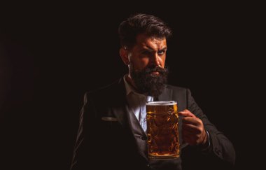 Serious man in classic suit drinking beer. Bearded guy in business outfit looks happy and satisfied. Portrait of man with lifted high glass of beer on black background.