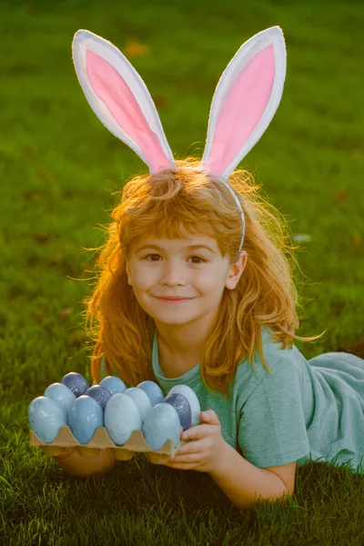 Kids boy in rabbit costume with bunny ears hunting easter eggs. Child gathering Easter eggs, laying on grass. — Stok fotoğraf