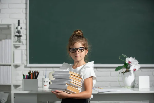 학교다니는 여자 애가 안경을 쓰고 칠판에 책을 쓴 채로요. 안경을 쓴 학교 아이는 칠판을 반대하는 심각 한 표현으로 책을 들고 있다. — 스톡 사진