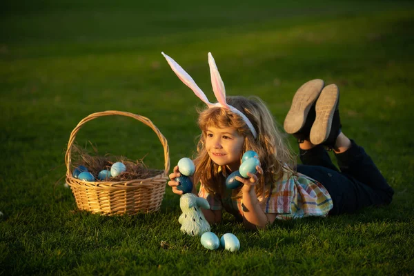 Child with easter eggs in basket outdoor. Boy laying on grass in park. Easter egg hunt. Fynny kids portrait. Child bunny boy with rabbit bunny ears. — Zdjęcie stockowe