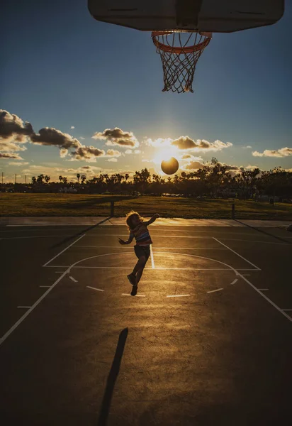 Dzieciak gra w koszykówkę. Cute little boy skoków dziecko z koszyka piłkę dla strzał sylwetka na zachód słońca. — Zdjęcie stockowe