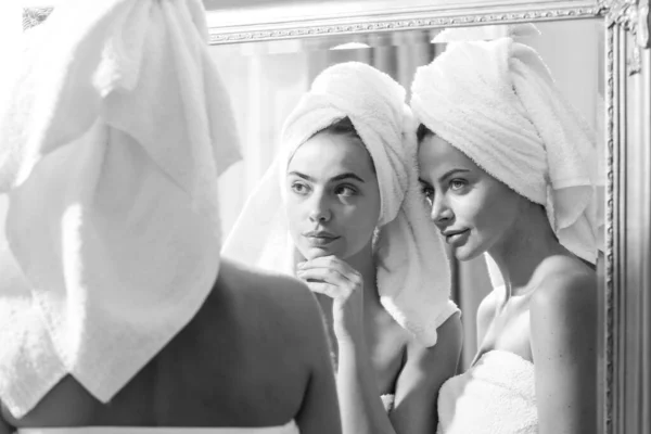 Mulher bonita olhar no espelho após o chuveiro no apartamento ou quarto de hotel. Jovens amigas meninas bonitas envolto em toalha alisamento aperfeiçoando a pele, conceito de rotina da manhã diária. — Fotografia de Stock
