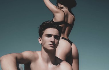 Beautiful young couple of lovers isolated at clear blue sky background. Sexy hot woman with fit sexy ass in bikini standing behind her handsome boyfriend. Sexy desire and love concept.