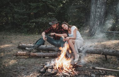 Beautiful young couple enjoying picnic in a park. Pleasant weekend near campfire. Couple in love on picnic or barbecue roasting marshmallows near bonfire. Friends sit on forest picnic.