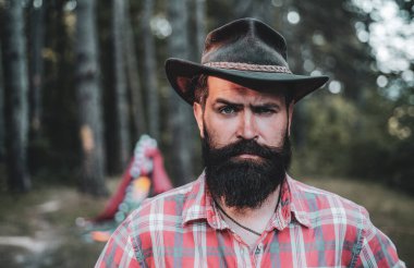 Mature hipster with beard. Brutal man with beard in hat. Brutal brunette bearded man in hat on a background of trees. Handsome strong stylish man with long lush black beard and moustache.