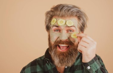 Yüzünde balçık maskesi ve salatalık dilimleri olan komik bir adam. Spa, cilt bakımı, sağlık ve yüz bakımı konsepti. Adamın kozmetik nemlendirici maskesi ve izole edilmiş bir stüdyo geçmişi var. Erkek yüz bakımı.