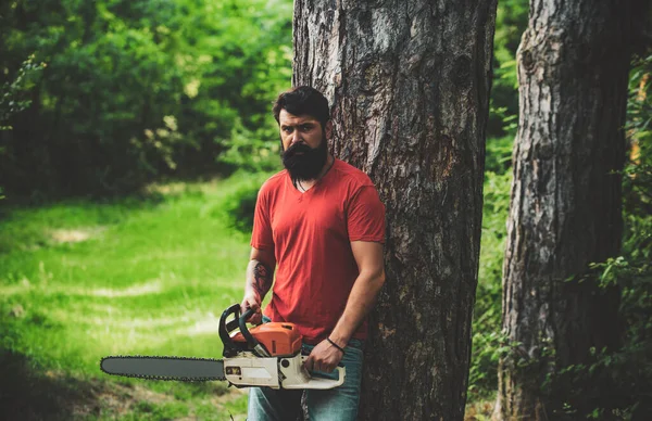 Holzfäller mit Kettensäge im Wald. Schöner junger Mann mit Axt in Waldnähe. Stilvoller junger Mann posiert wie ein Holzfäller. Ein schöner junger Mann mit Bart trägt einen Baum. — Stockfoto