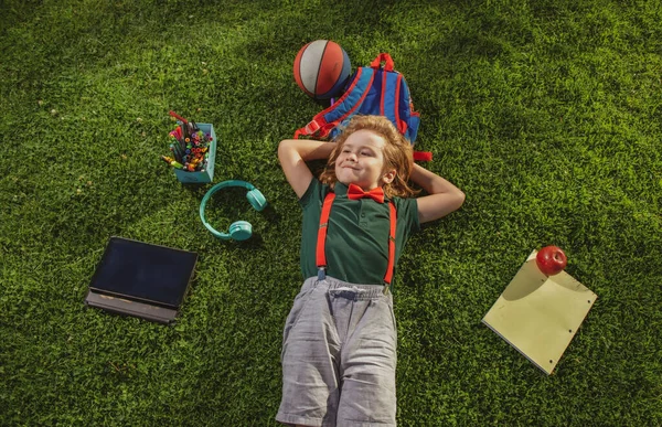 Schuljunge in der Natur. Nerd-Schüler mit Schulsachen Kind liegt im Gras. Traum vom Schulbesuch. Blick von oben auf kleinen Jungen, der auf grünem Gras liegt. — Stockfoto