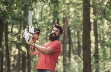 Ormanda elektrikli testereyle çalışan oduncu işçisi. Oduncu işçisi elektrikli testereyle ormanda yürüyor. Ormansızlaşma. Sakallı yakışıklı bir genç adam ağaç taşıyor..