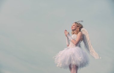 Charming curly little girl in white dress and wings - angel cupid girl. Portrait of a cupid little girl pray. Cherub. Cute teen cupid on the cloud - heaven background. Sweet angel girl.