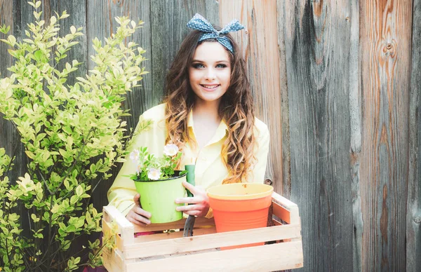 Pěkně usměvavá dívka drží zahradní nářadí na dřevěném pozadí. Koncept výsadby a pěstování květin. Zahradnice se stará o svůj dvorek. Rostliny jsou pojetí přátel. — Stock fotografie