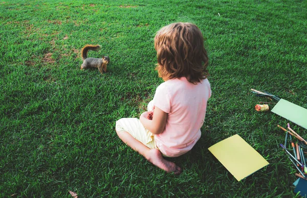 Parkta çimenlikte oturan çocuk öğrenci sincapla oynuyor. Çalış ve dışarıda öğren. Çocuk dışarıda okula hazırlanıyor.. — Stok fotoğraf