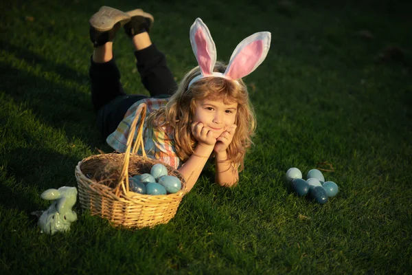 Enfant lapin mignon avec des oreilles de lapin. Enfant avec oeufs de Pâques dans le panier en plein air. Un gamin étendu sur l'herbe dans le parc. Chasse aux œufs de Pâques. Fynny enfants visage. — Photo