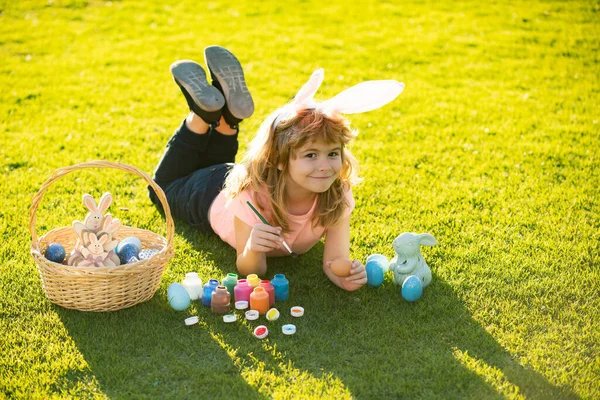 Kind jongen met paaseieren en konijnenoren leggen op gras schilderen eieren. — Stockfoto