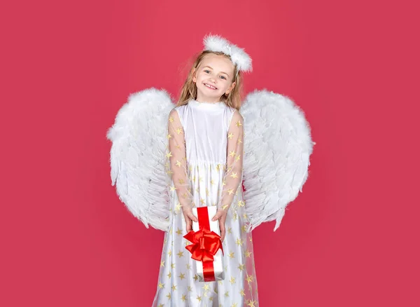 ¡Valentine! Pequeña chica Cupida. Ángel niño con regalo. Un angelito con alas blancas sostiene el regalo. Linda niña ángel con alas de ángeles, aislado en rojo. Tarjeta de regalo Valentine. — Foto de Stock