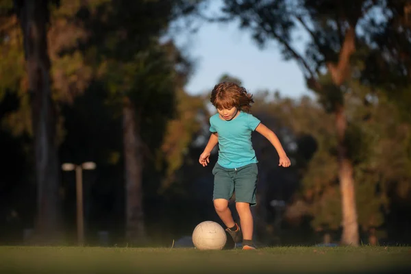 Garoto animado chutando bola na grama ao ar livre. Crianças de futebol, crianças jogam futebol. Criança jogando futebol no parque infantil no parque. — Fotografia de Stock