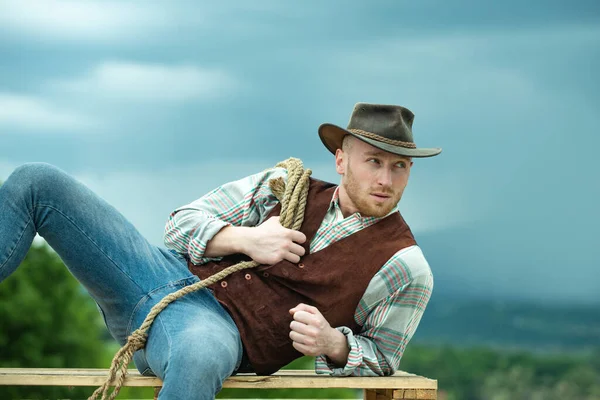 Ковбой на ранчо. Красивый мужчина в ковбойской шляпе и ретро-винтажном костюме. Ковбой с лассо-веревкой на фоне неба. — стоковое фото