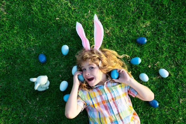 stock image Easter bunny children with rabbit ears in garden hunt egg. Child hunting eggs, wear bunny ears hold Easter eggs. Top view funny kids face.