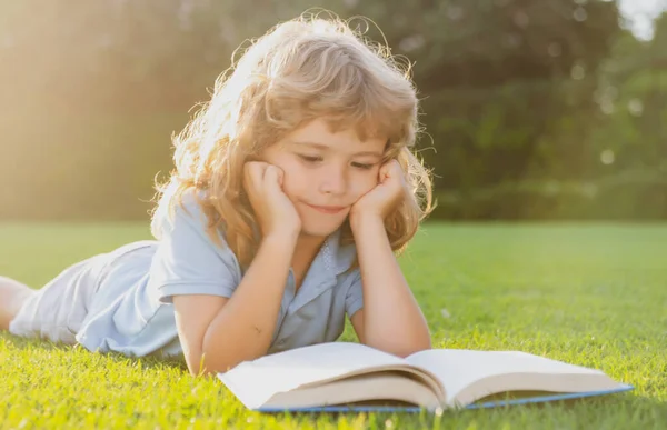 Niño leyendo un libro tirado en la hierba. Lindo niño pequeño con ropa casual leyendo un libro y sonriendo mientras está acostado en el césped en el parque. Concepto de aprendizaje, estudio, escuela y atención infantil. — Foto de Stock
