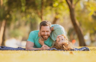 Baba ve oğul doğa parkında çimlerin üzerinde yatıyor. İnsanlar dışarıda eğleniyor. Mutlu tatiller ve dost canlısı aile kavramı.