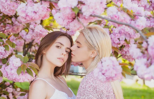 Νεαρά κορίτσια και ανοιξιάτικα λουλούδια. Πορτρέτο δύο όμορφων νεαρών γυναικών που χαλαρώνουν στα λουλούδια της σακούρα. Λεσβιακό ζευγάρι φιλιέται. Αισθησιακό άγγιγμα και φιλί. Ζευγάρι φιλενάδων. — Φωτογραφία Αρχείου