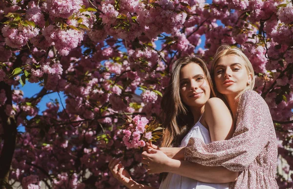 Dziewczyny w kwiatach. Odkryty portret dwóch młodych pięknych modnych kobiet pozujących w pobliżu kwitnącego drzewa sakura. Wiosenne dodatki i ubrania. Kobiece piękno i moda. Styl życia miasta. — Zdjęcie stockowe