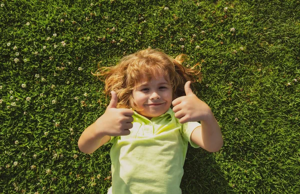 Grappig kind met duimen omhoog teken liggend op gras, dicht gezicht. Kinderen genieten van de zomer. — Stockfoto