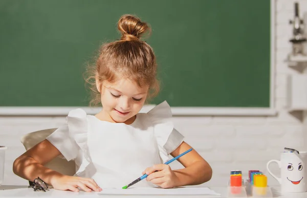 Kind meisje tekent in de klas zitten aan een tafel, plezier op school schoolbord achtergrond. kind leerling leren schilderen. — Stockfoto