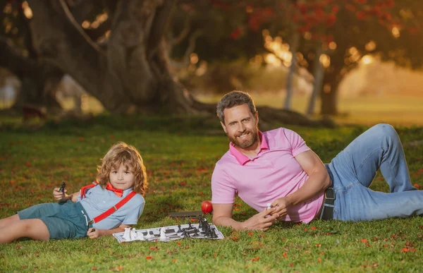 Fils étendu sur l'herbe et jouant aux échecs avec son père. Jeux et animations pour enfants. Concept de famille. — Photo