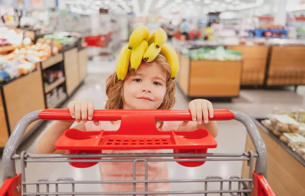 Banana on funny child head. Funny kid with shopping cart buying food at grocery store or supermarket. Surprised kids face. — Stock Photo, Image
