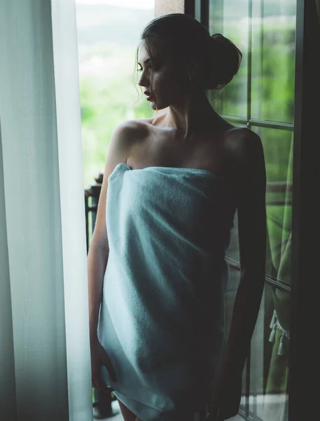 Zmysłowa młoda kobieta owinięta w ręcznik po prysznicu stojącym przy oknie i wyglądającym na zewnątrz. Piękna kobieta z luksusowym makijażem i fryzurą w tle okna. — Zdjęcie stockowe