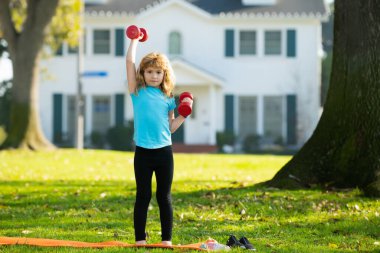 Dambıl kaldıran çocuk. Dambılı şirin çocuk eğitimi. Çocuk fitness. Çocuk dışarıda halterlerle egzersiz yapıyor. Sağlıklı çocukluk yaşam tarzı.