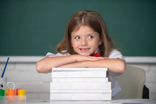 Wracać do szkoły. Uśmiechnięta dziewczyna siedząca za biurkiem i trzymająca się za ręce za książki w klasie szkolnej. — Zdjęcie stockowe