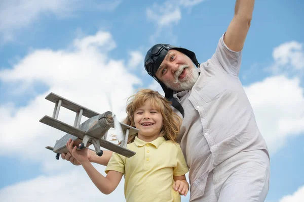 재밌는 손자와 할아버지가 여름 하늘을 배경으로 나무 비행기를 가지고 놀고 있다. 하늘을 나거나 여행하고 싶은 꿈을 가진 어린이. — 스톡 사진