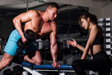 Spor salonunda dambılla egzersiz yapan bir sporcu. Kas yığını olan bir adamla kadın çalıştırıcı. İşlevsel antrenman yapıyor. Halteri kaldırıyor..