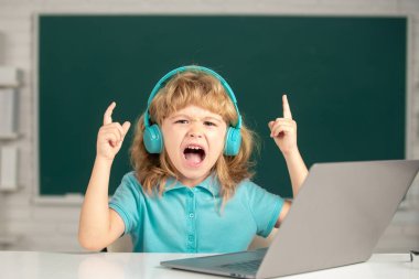 Kulaklıklı kızgın çocuk masada oturur, okulda dizüstü bilgisayarda çalışır, klever çocuk internet derslerini kullanarak öğrenirken kulaklık takar. Heyecanlı çocuk duyguları..