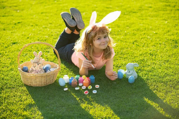 Kind met paaseieren en konijnenoren die op gras eieren verven. Gelukkige Pasen kinderen gezicht. — Stockfoto