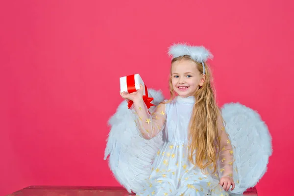 Engelenkinderen, gelukkige fee met cadeau, studioportret. Kleine blonde engel met witte vleugels heeft een geschenk. Schattig engelenmeisje met engelenvleugels, geïsoleerd op rood. Valentijnsdag. — Stockfoto