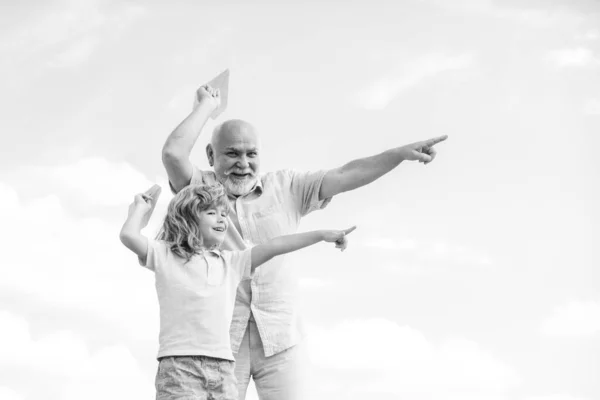 Junger Enkel und alter Opa spielen mit Spielzeugpapierflugzeug vor sommerlichem Himmel. — Stockfoto