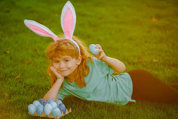 Lindo niño en orejas de conejo el día de Pascua. El niño busca huevos de Pascua en el césped. Niño con huevos de Pascua y orejas de conejo en la hierba en los rayos soleados. —  Fotos de Stock