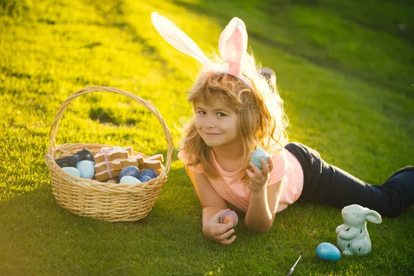 Chłopiec w stroju królika z króliczymi uszami polujący na wielkanocne jaja na trawie w parku wiosennym. Zabawny chłopiec, króliczki wielkanocne. — Zdjęcie stockowe